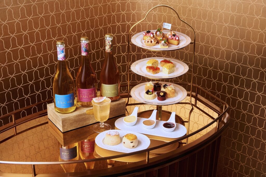 Three-tier afternoon tea stand with pastries and scones beside three bottles of Saicho sparkling tea on a brass trolley